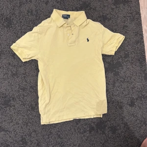 Gul pikétröja från Polo Ralph Lauren - Klassisk gul pikétröja från Polo Ralph Lauren med broderad marinblå logga på bröstet. Tröjan har korta ärmar, krage och knappar framtill. Tillverkad i bomull för en skön och luftig känsla.