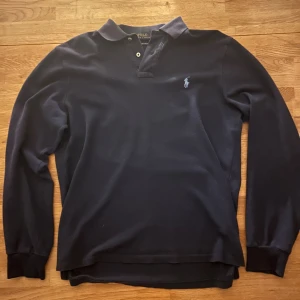 Marinblå pikétröja från Ralph Lauren - Snygg marinblå långärmad pikétröja från Polo Ralph Lauren i bomull. Tröjan har klassisk krage, två knappar vid halsen och den ikoniska broderade loggan på bröstet. Slim fit-modell som sitter riktigt schysst. Perfekt för dig som gillar stilrena och tidlösa plagg.