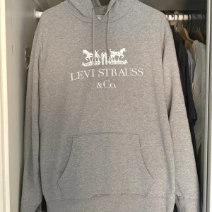 Grå hoodie från Levi's med tryck - Säljer en grå hoodie från Levi's med klassiskt vitt broderat tryck framtill. Tröjan har huva med snörning, stor magficka och långa ärmar. Perfekt för dig som gillar en enkel och stilren look med streetkänsla.