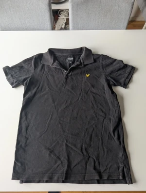 Svart pikétröja från Lyle & Scott - Svart pikétröja från Lyle & Scott med klassisk krage och korta ärmar. Tröjan har en gul broderad logga på bröstet och är tillverkad i mjuk bomull. Perfekt för en clean och enkel stil.