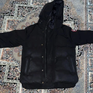 Svart dunjacka från Canada Goose - Svart dunjacka från Canada Goose med huva och klassisk logga på ärmen. Jackan har dragkedja och knappar framtill, två stora fickor och en quiltad design. Perfekt för kalla dagar med sin tjocka och varma fyllning. Snygg och stilren look som funkar till allt.