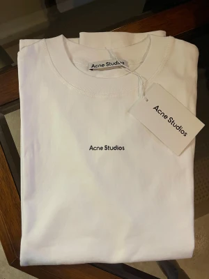 Vit Acne Studios t-shirt i bomull - Clean vit t-shirt från Acne Studios i 100% bomull. Klassisk passform med korta ärmar och rund halsringning. Diskret svart Acne Studios-logga broderad på bröstet. Perfekt basic-plagg för en stilren look.