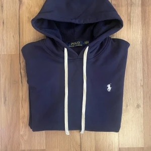Marinblå hoodie från Polo Ralph Lauren - Snygg mörkblå hoodie från Polo Ralph Lauren – helt ny med taggar. Tidlös design med vit broderad logga på bröstet. Perfekt basplagg för alla tillfällen.  💰 Pris: 649kr 📦 Fraktas snabbt