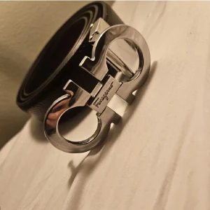 Svart Ferragamo bälte med silverdetaljer - Snyggt svart bälte från Ferragamo med ikoniskt dubbel-Gancini spänne i blankt silver. Bältet är tillverkat i äkta skinn och har en stilren, klassisk form som passar till jeans eller kostym. Spännet har Ferragamo-loggan ingraverad och ger en lyxig vibe.