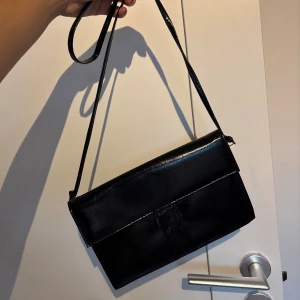 Svart axelväska i skinn - Snygg svart axelväska i glansigt material med stilren, rektangulär form och smal axelrem. 