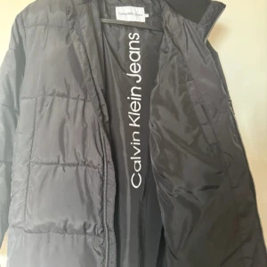 Svart pufferjacka från Calvin Klein Jeans - Svart pufferjacka från Calvin Klein Jeans med huva och dragkedja. Jackan har ett stilrent CK Jeans-märke på ärmen och stort logotryck på insidan. Lång modell med quiltad design och skön passform, perfekt för kalla dagar.