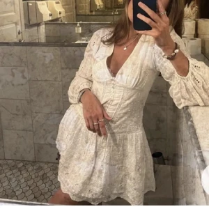 Vit kort klänning med spetsdetaljer student klänning - Supersöt vit kort klänning med v-ringning och spetsdetaljer. Klänningen har puffiga långärmar, markerad midja och ett flowigt kjolparti. Perfekt för sommarens alla tillfällen och ger en romantisk vibe. Materialet är lätt och luftigt bomull med broderade detaljer.