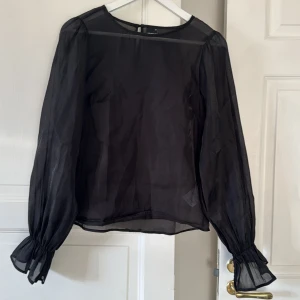 Svart transparent blus från Gina Tricot - Säljer en svart transparent blus från Gina Tricot i storlek 38. Blusen har långa puffärmar med rynkade muddar och rund halsringning. Stängs med en liten knapp i nacken. Perfekt för dig som gillar en stilren och trendig look.