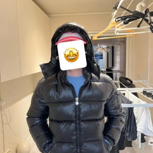 Svart Moncler pufferjacka med huva - Säljer en svart pufferjacka från Moncler med glansig finish och stor, vadderad huva. Jackan har dragkedja framtill, sidofickor och Moncler-logga på ärmen. Perfekt för kalla dagar och har en riktigt schysst passform. Jackan är använd och små slitage på insaidan som inte syns när du har på den. Det ända som är lite skavt är ett litet hål på ryggen som man kan se på bilden. 