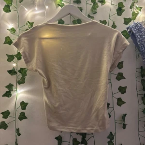 Beige croppad topp med öppen rygg - Säljer en beige croppad topp med korta ärmar och en stor öppen rygg. Toppen har en enkel och clean look framtill och ett elastiskt band nedtill på ryggen för en snygg passform. Perfekt för dig som gillar minimalistisk stil.