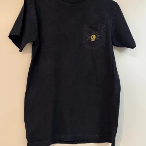 Svart t-shirt Welcome S - Svart t-shirt från Welcome i 100% bomull med coolt tryck i guld på bröstfickan och ryggen. Trycket föreställer en kristallkula med måne och strålar samt texten 'WELCOME OBSCURUS NAVITAS'. Klassisk passform och rund hals. Slitage krage, se sista bilden.