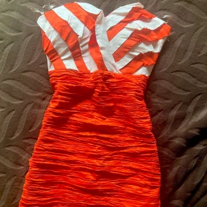 Röd och vit randig kortklänning - Säljer en snygg kortklänning med röd och vit randig överdel och röd rynkad kjol. Klänningen är axelbandslös med hjärtformad urringning och har en tight passform. Materialet känns som satin eller polyester och ger en cool struktur. Perfekt för dig som vill sticka ut med färg och mönster.