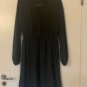 Svart långärmad klänning med knappar VeroModa - Svart klänning med lång ärm och knappar framtill. Klänningen har en markerad midja och ett enkelt, stilrent snitt som ger en klassisk look. Perfekt för dig som gillar minimalistisk stil och vill ha något som funkar till många olika tillfällen.