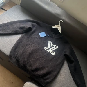 Svart stickad Louis Vuitton tröja - Svart stickad tröja från Louis Vuitton med stort vitt LV-logo framtill. Rund hals och ribbade muddar vid ärmslut och nederkant. Perfekt för dig som gillar exklusiva märken och vill ha en clean look med tydlig branding.