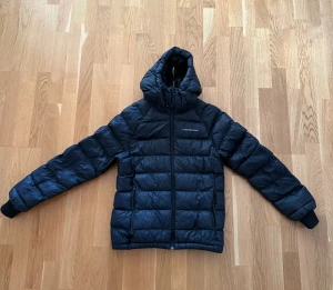 Svart dunjacka från Peak Performance - Säljer en svart dunjacka från Peak Performance med huva och dragkedja framtill. Jackan har quiltad design och långa ärmar med muddar. Perfekt för kalla dagar och har en stilren look som funkar till det mesta. 