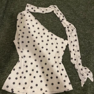 Prickig vit oneshoulder topp med scarf - Säljer en vit topp med svarta prickar i oneshoulder-modell. Toppen har en insydd scarf som går att designa på olika vis. Säljer då den aldrig kommit till användning.