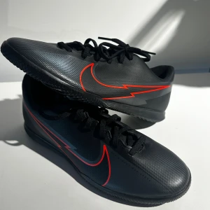 Nike fotbollsskor svart/röd strl 40 - Svarta Nike fotbollsskor med röd blixtformad Swoosh på sidan. Skorna har snörning och en platt, mönstrad sula för bra grepp på planen. Tillverkade i syntetmaterial med låg profil och rund tå. Perfekt för dig som vill sticka ut med stil på fotbollsplanen.