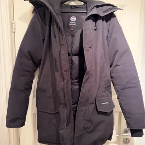 Svart parkas från Canada Goose - Äkta Canada Goose Langford Parka (M). Varm parkas (-15 C till -25 C), svart. Bra skick, sparsamt använd. OBS: Litet hål vänster underarm (se bild). Alla funktioner, dragkedjor, muddar i bra skick. Avtagbar äkta coyotepäls. Äktheten garanteras med hologrametikett (se bild). Originalkvitto saknas. Pris diskuteras. Fler bilder finns!
