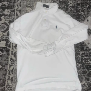 Vit långärmad piké från Polo Ralph Lauren - Vit långärmad pikétröja från Polo Ralph Lauren med klassisk krage och knappar framtill. Ikoniska broderade loggan i mörkblått på bröstet. Skön och stilren, perfekt till jeans eller chinos.