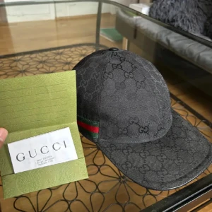 Svart keps från Gucci med GG-mönster - Säljer en svart keps från Gucci med klassiskt GG-monogram över hela kepsen. Kepsen har en böjd skärm, justerbart spänne bak och en ikonisk grön-röd rand på sidan. Tillverkad i bomullscanvas med läderdetaljer. Perfekt för dig som vill ha en lyxig streetstyle-look.