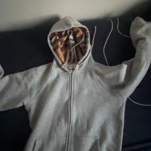 Grå hoodie från Burberry med dragkedja - Säljer en grå hoodie från Burberry med klassiskt rutigt foder i huvan. Tröjan har dragkedja framtill, snören vid huvan och broderad logga på bröstet. Perfekt för dig som gillar stilrena och lyxiga streetwear-plagg.