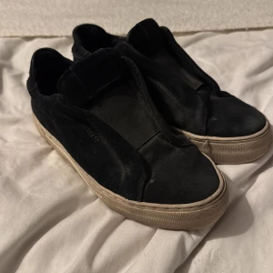 Svarta sneakers från Axel Arigato - Svarta sneakers från Axel Arigato med stilren design och bred resår över vristen. Ovandel i mocka och skinn, vit platt sula och diskret logga på plösen. Perfekta för dig som gillar minimalistisk och clean stil.