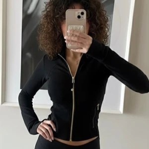 Svart träningsjacka från Lululemon - Svart figursydd träningsjacka från Lululemon med guldiga dragkedjor och två fickor framtill. Jackan har lång ärm, hög krage och är tillverkad i ett stretchigt material som passar perfekt till aktiva dagar.