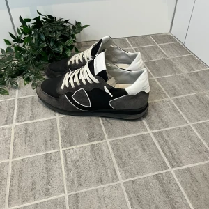 Philippe Model svarta sneakers i mocka - Snygga sneakers från Philippe Model i svart och grå mocka med vita detaljer och snörning. Skorna har en klassisk rund tå och platt sula, samt vit hälkappa och vita skosnören. Perfekta för dig som gillar stilrena och trendiga sneakers.