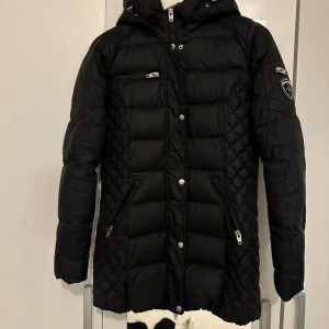 Svart dunjacka Rockandblue med huva - Svart dunjacka från Rockandblue med huva, quiltade detaljer och flera dragkedjefickor. Jackan har en längre modell, snygga silvriga knappar och Rockandblue-logga på ärmen. Perfekt för kalla dagar och riktigt stilren look.