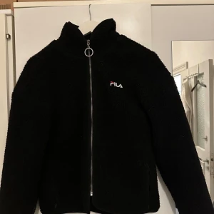 Svart teddyjacka från FILA - Svart teddyjacka från FILA med stor logga på ryggen och mindre logga på bröstet. Jackan har hög krage, hel dragkedja med rund zip puller och är tillverkad i mjukt fleece-material. Perfekt för kalla dagar och har en clean, sportig vibe.