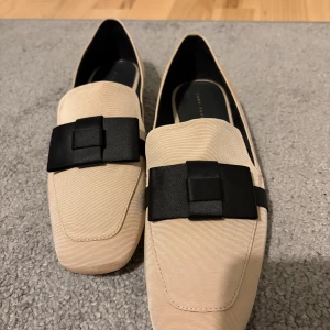 Beige loafers med svart rosett Zara - Stilrena beige loafers från Zara Trafaluc med fyrkantig tå och dekorativ svart rosett framtill. Skorna har låg klack och är tillverkade i ett tygmaterial som ger en elegant känsla. Perfekta för dig som gillar klassisk men trendig stil.