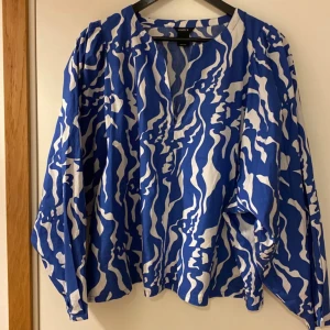 Blåvit mönstrad blus från Lindex large  - Säljer en blå och vit blus från Lindex med vågigt mönster. Blusen har lång ärm, v-ringning och en lös passform. Perfekt för dig som gillar färg och mönster i garderoben.