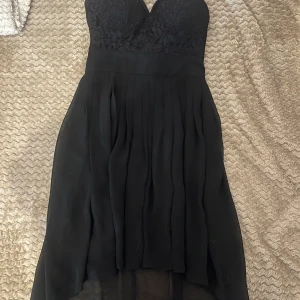 Svart spetsklänning med v-ringning - Supersnygg svart klänning med v-ringad överdel i spets och plisserad kjol. Klänningen är ärmlös och har en elegant, asymmetrisk fåll som är längre bak. Perfekt för dig som vill sticka ut med en klassisk men ändå modern look.
