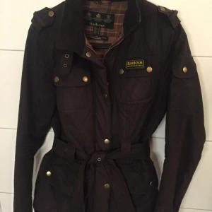 Barbour jacka  - Snygg brun jacka från Barbour med klassisk tartanrutig insida. Jackan har flera fickor med knappar, dragkedja och bälte i midjan. Perfekt för dig som gillar en tidlös look med coola detaljer.