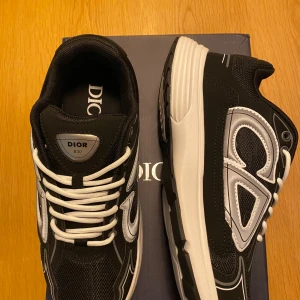 Svarta Dior B30 sneakers - Säljer ett par svarta Dior B30 sneakers med vita och silverfärgade detaljer. Skorna har en chunky sula, snörning och meshpartier för extra ventilation. Snyggt Dior-märke på sidan och tungan. Perfekt för dig som gillar exklusiva och trendiga sneakers.