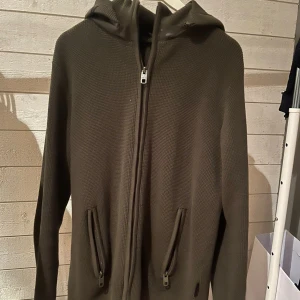 Olivgrön hoodie med dragkedja - Säljer en olivgrön hoodie med huva och hel dragkedja framtill. Tröjan har två fickor med dragkedja och är tillverkad i ett grovstickat material. Perfekt för dig som gillar en chill och enkel stil. Nästintill aldrig använd