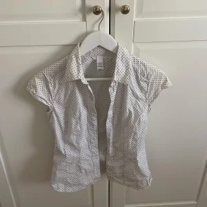 Vit kortärmad prickig topp från H&M - Snygg vit skjorta från H&M med svarta prickar, korta ärmar och klassisk krage. Skjortan har knäppning framtill och är gjord i ett lätt bomullstyg, perfekt för en fräsch och stilren look. Men används även som en skit snygg fest topp. Den kan även knäppas.