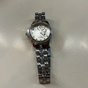 Silverfärgad klocka från Geneva - Stilren klocka från Geneva i silverfärgat metallarmband med dekorativa stenar runt urtavlan. Urtavlan har blommigt mönster och markerade siffror. Perfekt accessoar för att lyfta din outfit. 