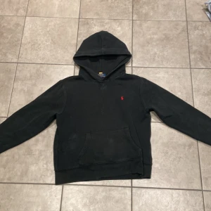 Svart hoodie från Polo Ralph Lauren - Svart hoodie från Polo Ralph Lauren med klassisk röd logobrodering på bröstet. Tröjan har huva, lång ärm och en stor magficka framtill. Mjuk och skön bomullskvalitet som passar perfekt när du vill ha en chill och stilren look.OBS den kan se mörkgrön ut i ljuset!