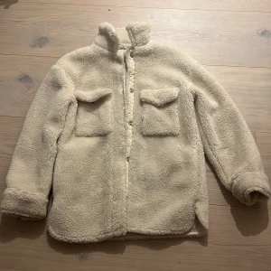 Vit barnteddyjacka med fickor - Mysig vit  barnteddyjacka med två stora bröstfickor och ståkrage. Jackan har knappstängning framtill och är tillverkad i fluffigt teddy-material som håller dig varm. Perfekt för dig som gillar en chill och trendig look under kyliga dagar.