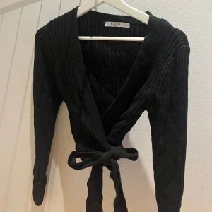 Svart omlottopp från NA-KD - Säljer en svart ribbad omlottopp från NA-KD med långa ärmar och knytband i midjan. Toppen har v-ringning och är croppad, vilket ger en snygg och trendig look. Perfekt att styla med höga jeans eller kjol.