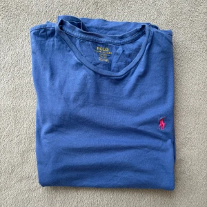 Blå t-shirt från Polo Ralph Lauren - Snygg blå t-shirt från Polo Ralph Lauren med klassisk röd broderad logga på bröstet. T-shirten har rund halsringning och är tillverkad i mjuk bomull. Perfekt för en clean och stilren look.