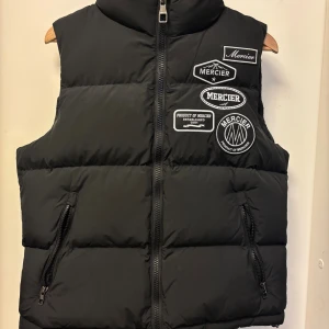 Svart dunväst från Mercier - Svart dunväst från Mercier med flera broderade patches på bröstet. Västen har två dragkedjefickor framtill och stängs med dragkedja. Klassisk puffer-modell med hög krage och diskret logga på ryggen. Perfekt för lager-på-lager-stil.