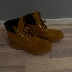 Timberland boots (mocka) - Klassiska Timberland boots i gulbrun mocka med svart vadderad kant upptill. Grov sula för bra grepp, snörning med guldfärgade öljetter och rund tå. Perfekta för höst och vinter, ikonisk logga på sidan och robust känsla. Använd 1 gång utomhus säljes pga fel storlek. Nypris 2400kr. 