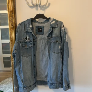 Blå jeansjacka med paisley från Catch - Oanvänd - Säljer en blå jeansjacka från Catch i storlek M. Jackan har slitna detaljer, knappar framtill och två bröstfickor. På ryggen finns ett stort paisley-mönster i svart som ger jackan en unik vibe. Perfekt för dig som gillar streetstyle och vill sticka ut.