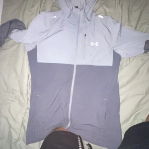 Grå vindjacka från Under Armour - Snygg tvåtonad grå vindjacka från Under Armour med huva och hel dragkedja framtill. Jackan har lång ärm, bröstficka med dragkedja och logga på bröstet. Perfekt för blåsiga dagar och sportiga outfits.