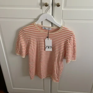 Randig t-shirt från Zara i L - Fräsch t-shirt från Zara i storlek L med smala orangea ränder på en krämvit botten. Klassisk rund hals och korta ärmar. Mjuk och skön bomullskvalitet som känns lätt mot huden. Perfekt för dig som gillar enkel men snygg stil. Den är helt oanvänd och passar till alla typer av jeans.