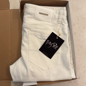 Vita slim jeans från Sacré Jeans - Slim fit jeans i fräsch vit färg från Sacré Jeans, modell SLIM/1111. Klassisk femficksdesign med metallnitar och knappstängning. Snyggt minimalistiskt utseende och raka ben, perfekta för en clean och modern look.