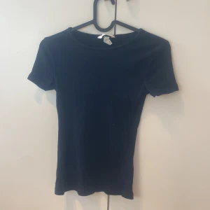 Svart ribbad topp från H&M XS - En svart ribbad topp från H&M i storlek XS. Toppen har korta ärmar, rund halsringning och en slim passform som sitter snyggt. Perfekt att matcha med jeans eller kjol för en clean look.