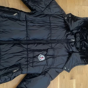 Svart Moncler dunjacka med huva - Säljer en svart dunjacka från Moncler med glansig finish, huva och dragkedja framtill. Jackan har två sidofickor med dragkedja och en bröstficka med Moncler-logga. Perfekt för kalla dagar och riktigt snygg streetstil.